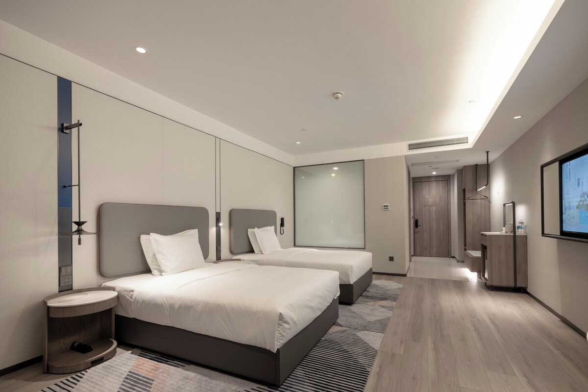 Una habitación de hotel moderna con dos camas, un espejo grande, un televisor y un baño con lavamanos y espejo.