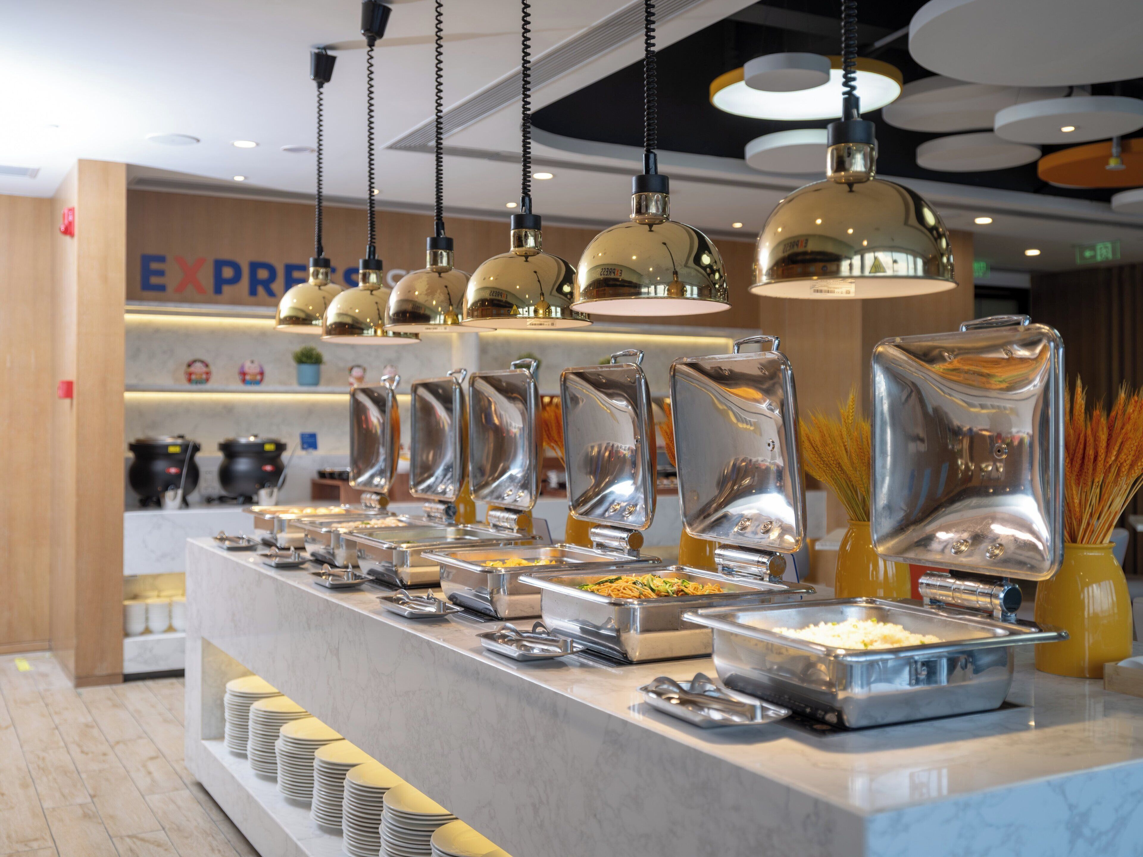 A buffet area with multiple food stations, stainless steel serving trays, and hanging pendant lights.