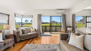 Amplia sala de estar con un gran ventanal que ofrece vistas a un campo de golf. Hay un sofá color beige, un sillón estampado y una mesita con una lámpara.