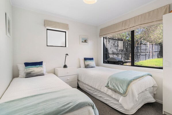 A bedroom with two beds, a nightstand, a lamp, a window with a view of a backyard, and a framed picture on the wall.
