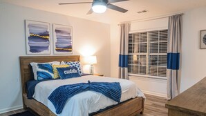 Un dormitorio con una cama de madera, una mesita de noche con lámpara, una ventana con persianas y dos cuadros abstractos en la pared.