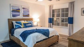 A bedroom with a wooden bed, a nightstand with a lamp, a window with blinds, and two abstract paintings on the wall.