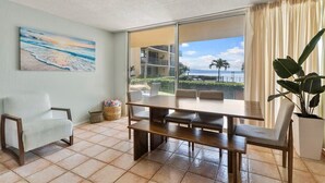Ein Zimmer mit einer großen Schiebetür, die auf einen Balkon mit Blick aufs Meer und Palmen führt. Dort steht ein Holztisch mit Stühlen, ein weißer Sessel und ein Blumentopf.