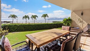 Eine Terrasse mit Holztisch und Korbstühlen, mit Blick auf einen Strand mit Palmen und einem klaren Himmel.