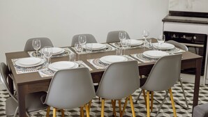 A dining area with a rectangular table set for six, surrounded by six grey chairs with wooden legs. The table is covered with a patterned table runner and set with white plates, glasses, and cutlery. The floor has a black and white checkered pattern, and a kitchen with a stove is visible in the background. - Lagarteiro House (Porto)