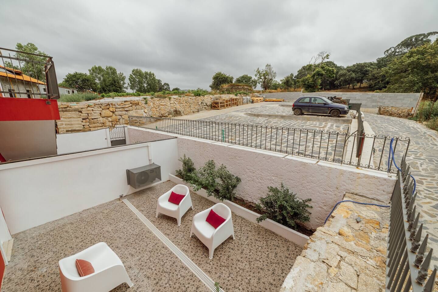 A rooftop terrace with stone flooring, white chairs with red cushions, a car, and a stone wall.