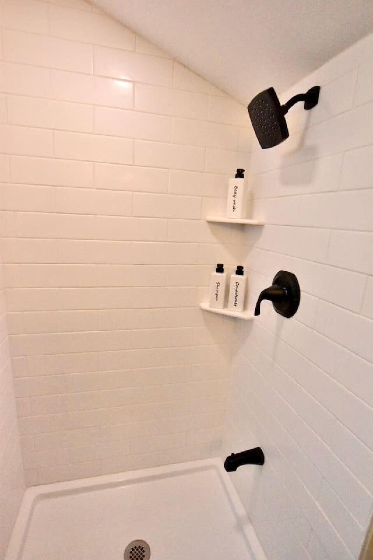 A shower area with white tiled walls, a black showerhead, and two white bottles on a shelf.