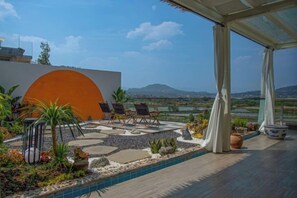A poolside area with a view of a mountain range, outdoor seating, and a large orange wall.