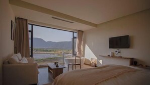 A bedroom with a bed, a desk, a chair, a TV, and a large window with a view of mountains.