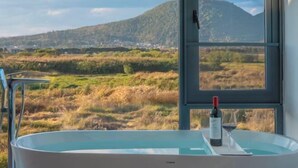 A bathtub with a bottle of wine and a glass, a window with a view of a mountainous landscape, and a metal faucet.