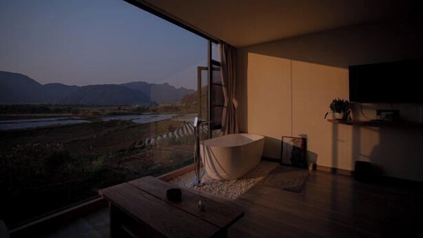 A modern bathroom with a bathtub, a wooden bench, and a large window offering a view of a river and mountains.