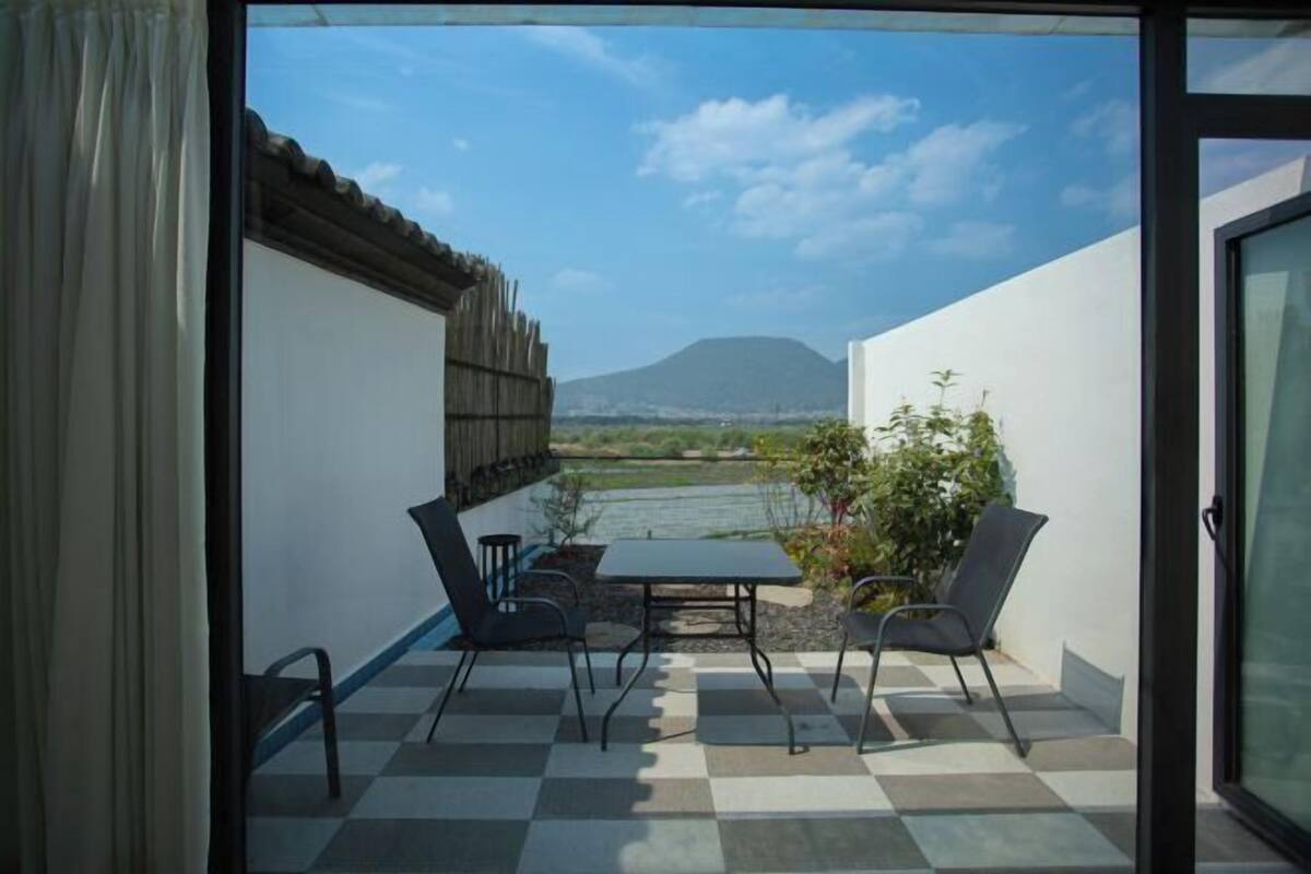 Pátio aberto com chão xadrez, mesa e cadeiras, com vista para um corpo d'água e uma montanha.