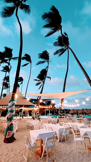An outdoor seating area with tables and chairs, palm trees, and string lights against a clear sky.