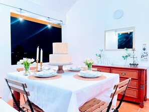 A dining area with a table set for two, featuring a cake centerpiece, white plates, and candles.