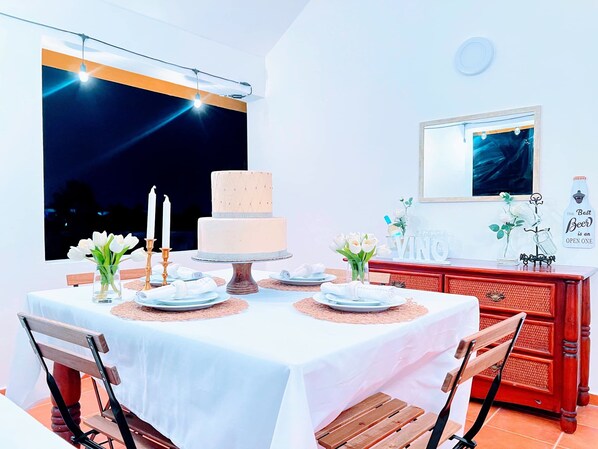 A dining area with a table set for two, featuring a cake centerpiece, white plates, and candles.