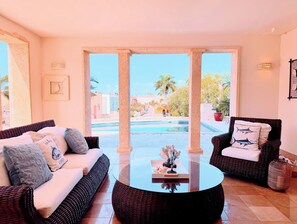 Ein Zimmer mit einem Korbssofa, einem runden Glastisch und Blick auf einen Swimmingpool.