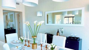 Un coin repas avec une longue table dressée pour quatre personnes, des chaises blanches et un grand miroir au-dessus.