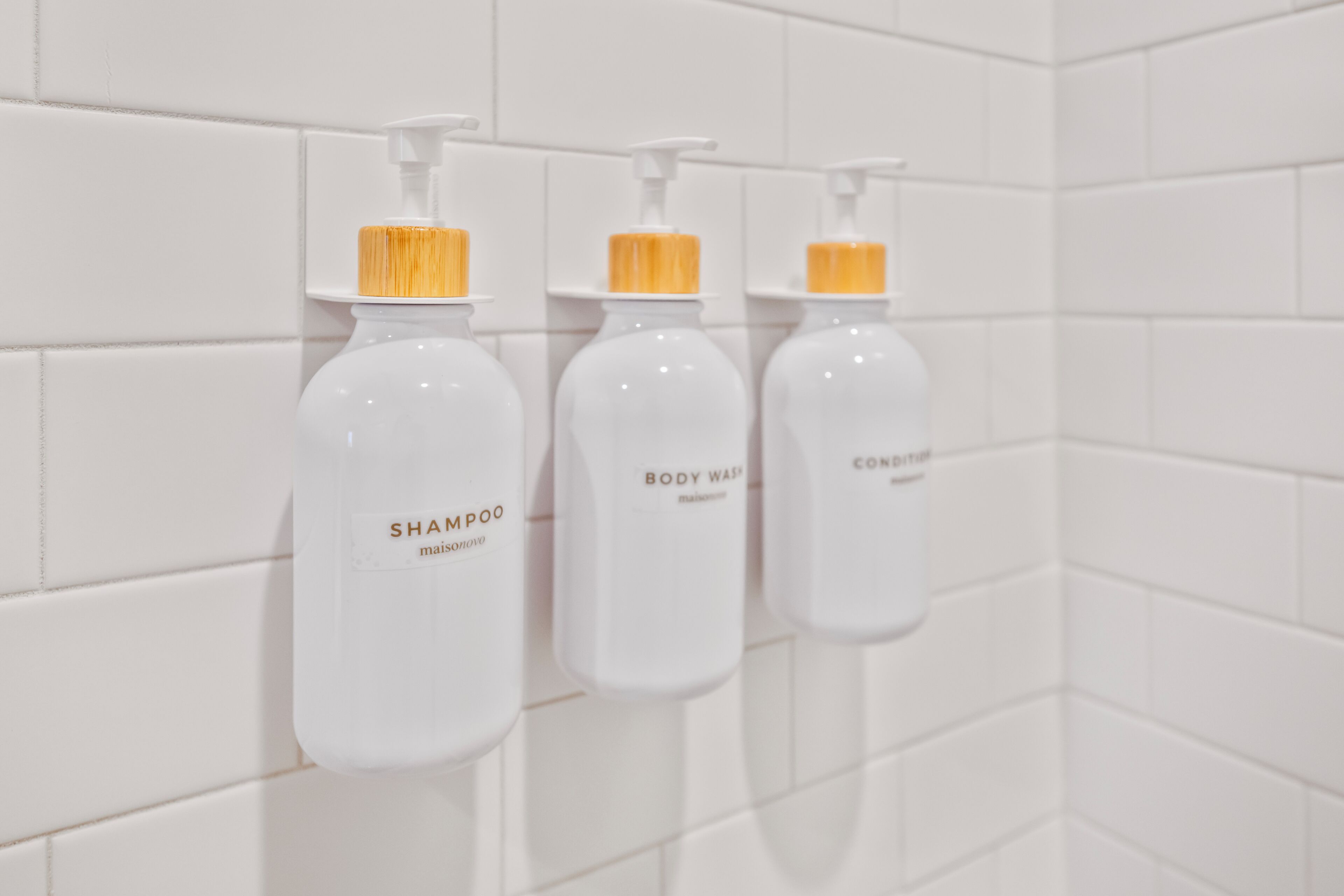 Tres botellas blancas con tapas amarillas etiquetadas 'SHAMPOO', 'BODY WASH' y 'CONDITIONER' montadas sobre una pared de azulejos blancos.