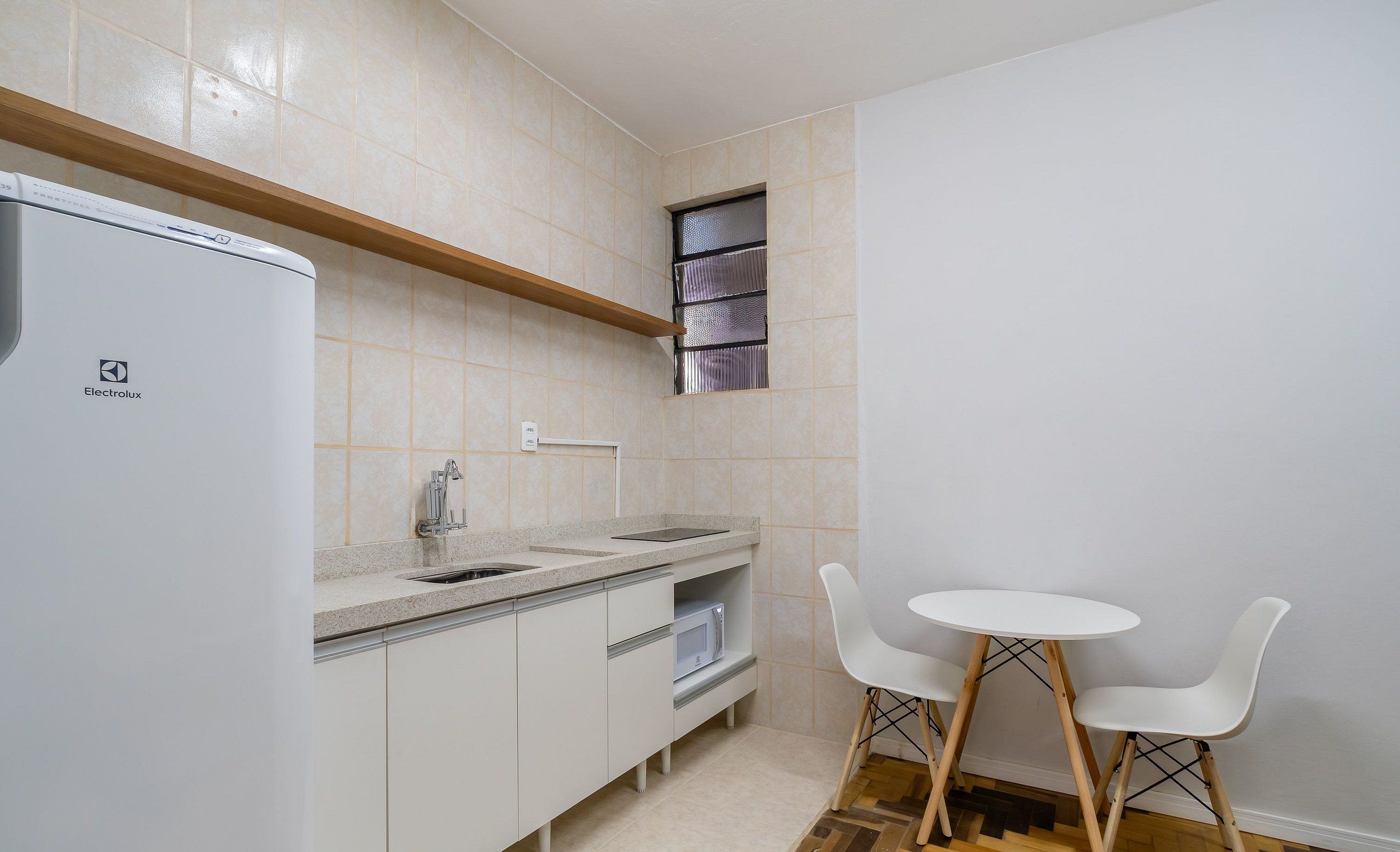 Een compacte keuken met een inbouwkoelkast, een spoelbak en een klein raam. Er staat een witte tafel met twee stoelen en de vloer heeft een visgraatmotief.