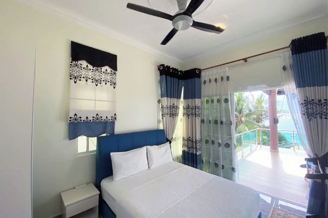 Un dormitorio con cama, mesita de noche, ventilador de techo y vistas a un balcón con cortinas.