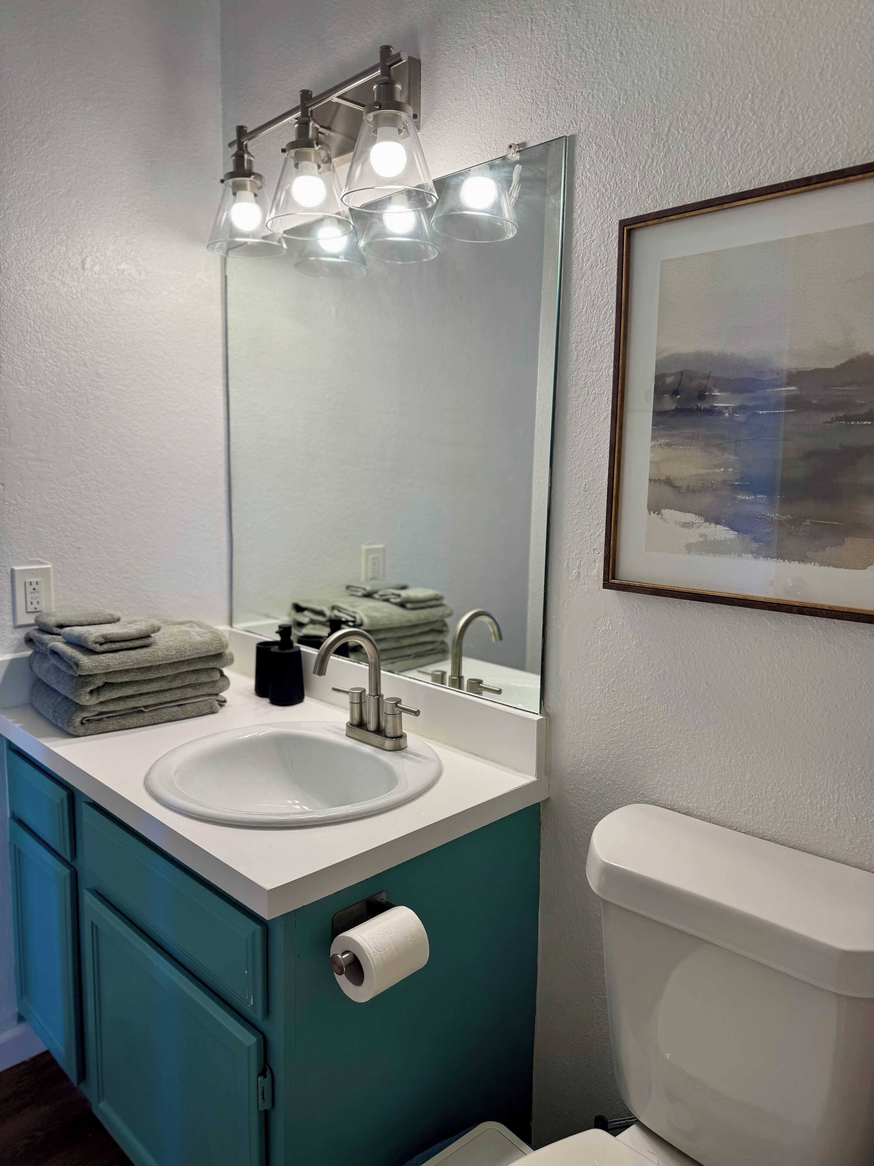 A bathroom with a white sink, a mirror, a toilet, teal cabinets, and a framed picture on the wall.