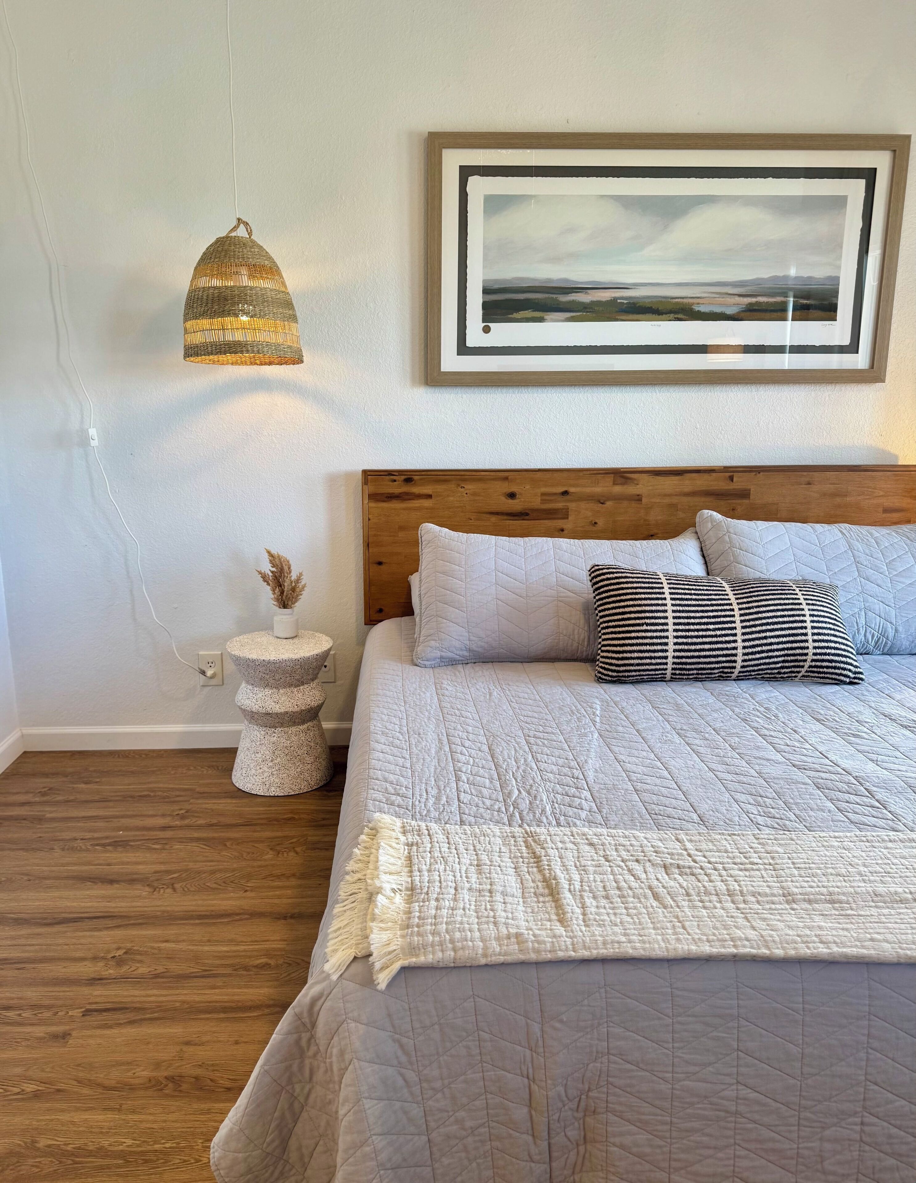 A bedroom with a wooden headboard, a large bed with multiple pillows, a hanging light fixture, and a framed landscape painting on the wall.