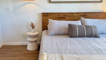 A bedroom with a wooden headboard, a large bed with multiple pillows, a hanging light fixture, and a framed landscape painting on the wall.