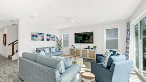 Una sala moderna con un sofá gris, un televisor montado en la pared y un amplio ventanal que deja entrar luz natural.