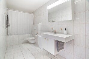 Un bany modern amb rajoles blanques, una banyera, un vàter, un lavabo amb mirall i un suport per a tovalloles.