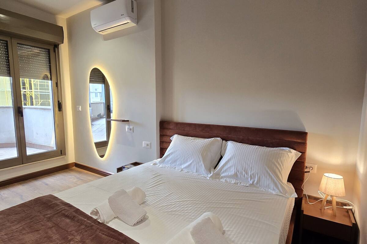 A hotel room with a large bed, a wooden headboard, a bedside table with a lamp, and a window with blinds.