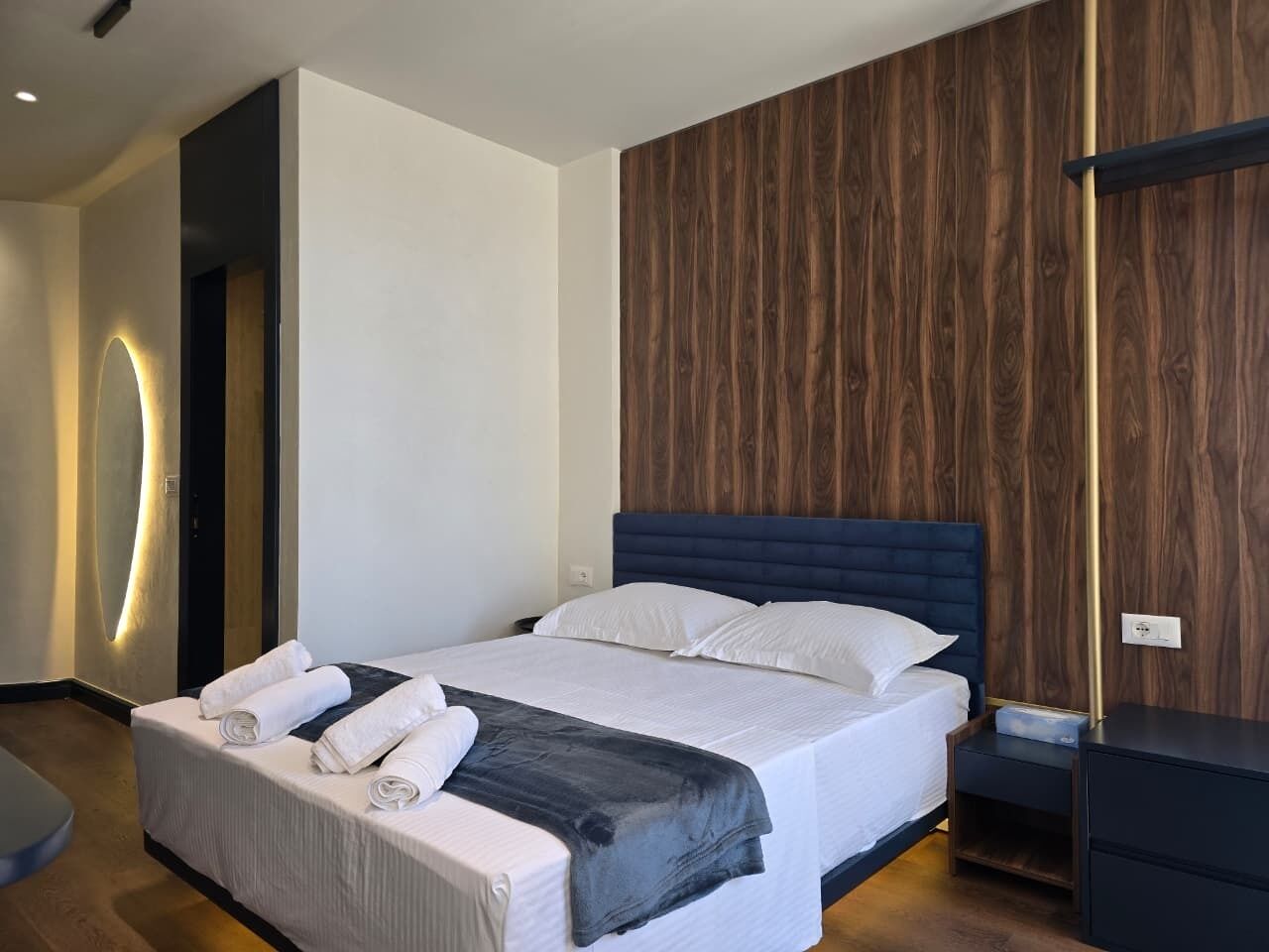 A modern hotel room with a large bed, a wooden accent wall, and a bedside table.