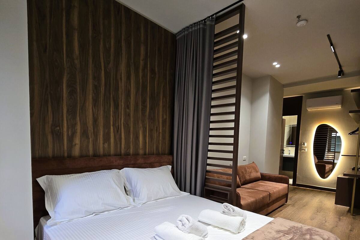 A modern hotel room with a large bed, a wooden headboard, a brown sofa, and a small seating area with a mirror and lighting.