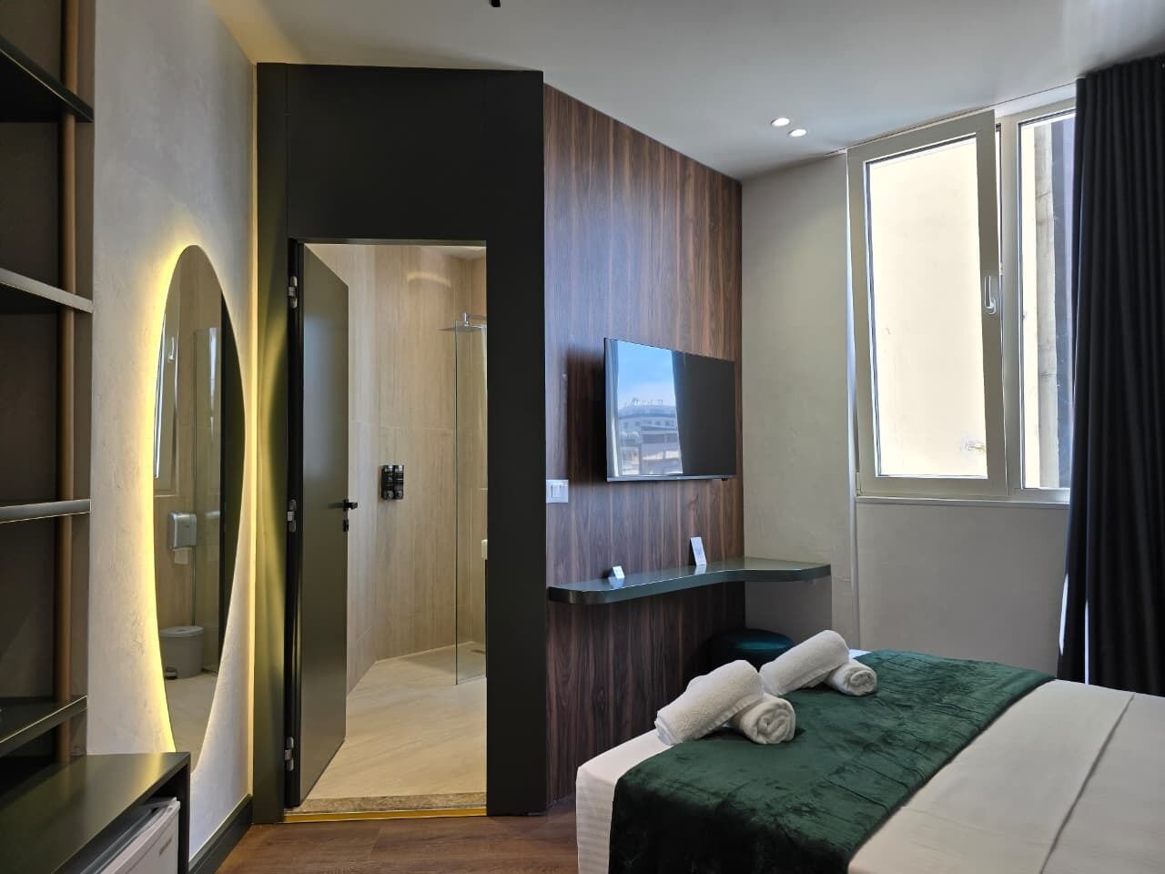 A modern hotel room with a bed, a TV, a mirror, and a bathroom visible through an open door.