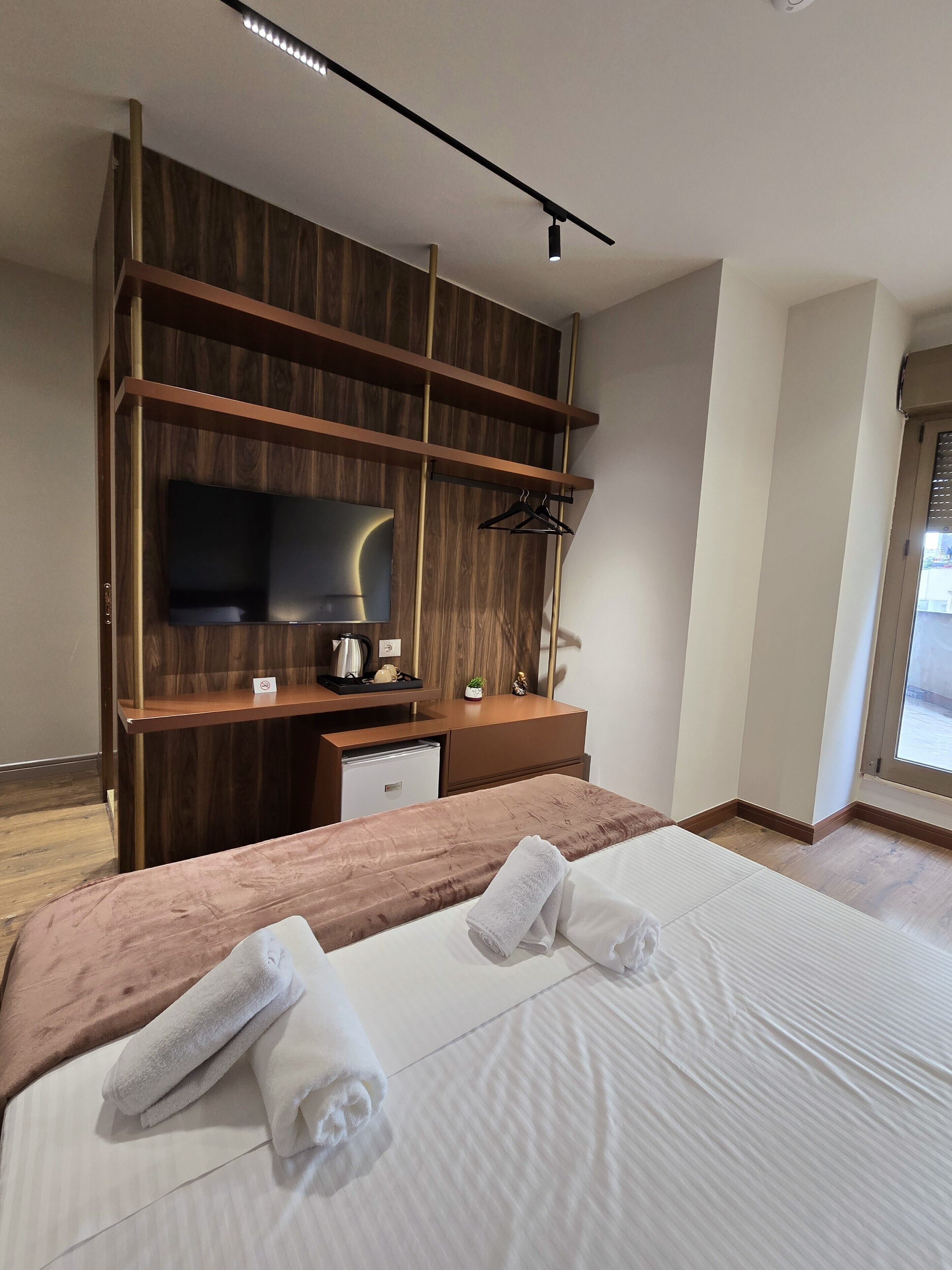 A modern hotel room with a large bed, a flat-screen TV mounted on a wooden panel wall, a small desk with a kettle and a cup, and a window with a view of the outside.