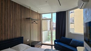 Habitación de hotel con cama, sofá, televisión y balcón con vista a edificios.