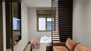 Una habitación moderna con suelo de madera, un sofá marrón, un televisor en la pared, una cama con colchón blanco y manta marrón, y una ventana con persianas.