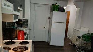 A compact kitchen with white cabinets, a white refrigerator, and a stove. There is a wooden chair and a small table with a lamp.