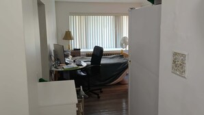 A narrow hallway leading to a bedroom with a bed covered by a green sheet. A desk with a computer and chair is visible in the room.