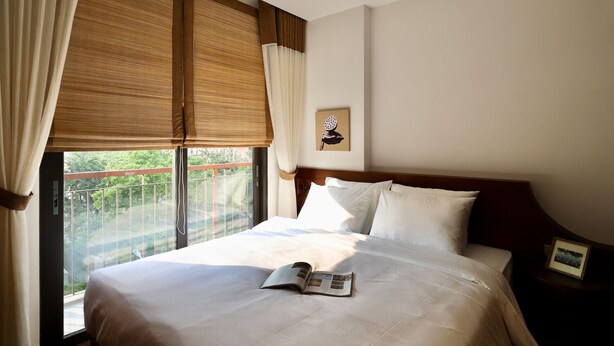 Una habitación de hotel con una cama grande, una ventana con cortinas, un cuadro en la pared y una mesita de noche con una lámpara.