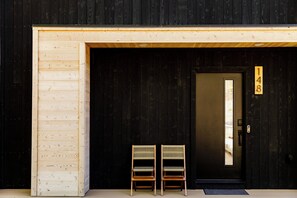A wooden door with a number plaque, two wooden chairs outside, and a dark wooden wall.