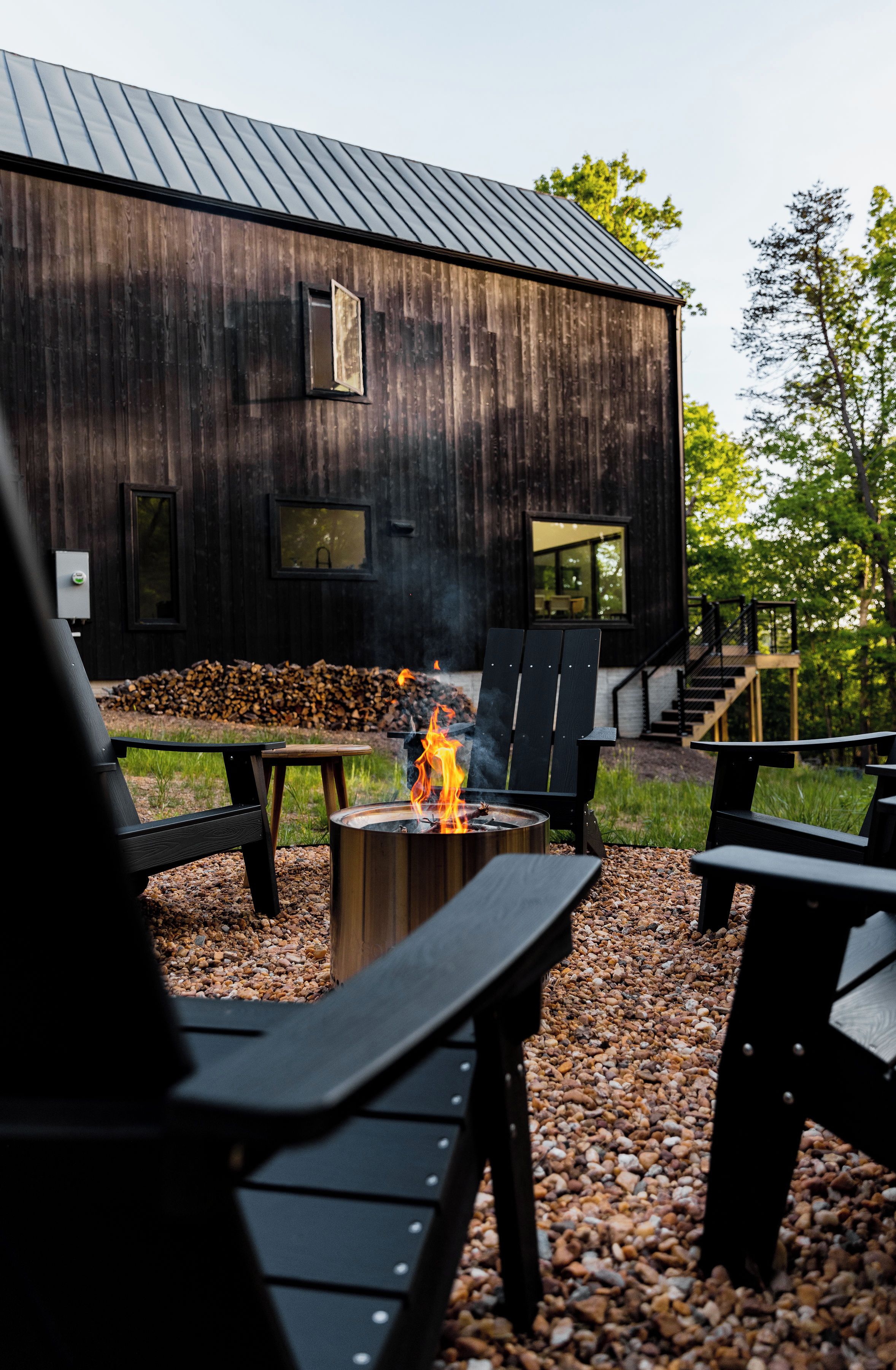 Un espace extérieur aménagé dans un style rustique, avec des fauteuils Adirondack, un foyer et un bâtiment en bois doté de grandes fenêtres.
