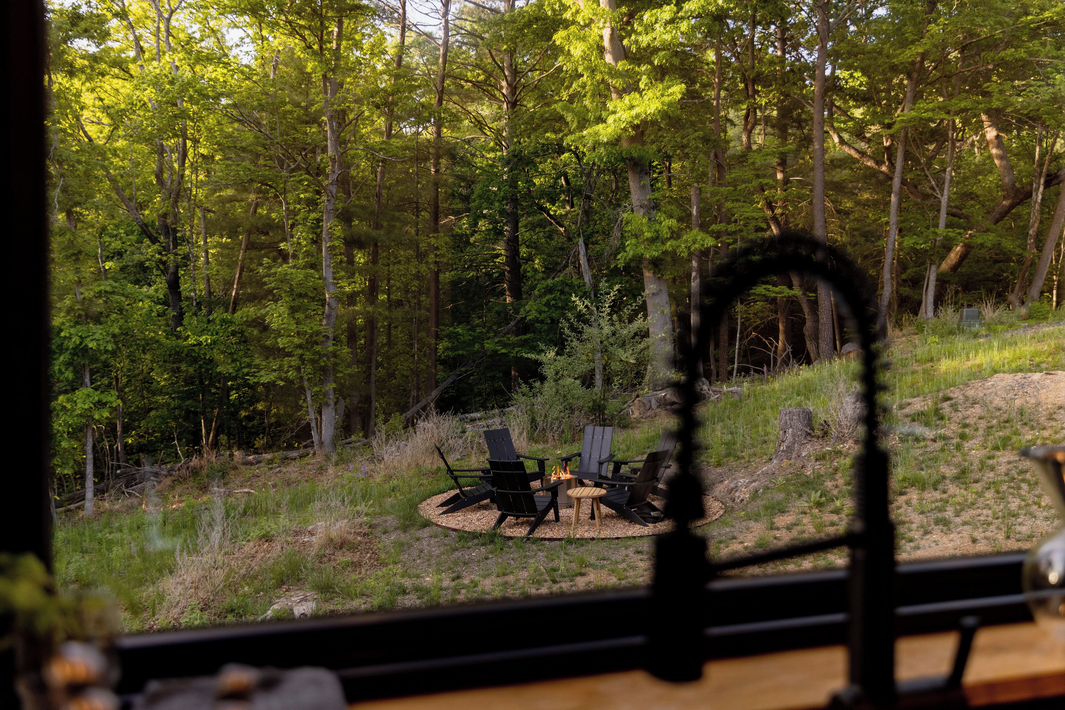A forested area with a fire pit and chairs.