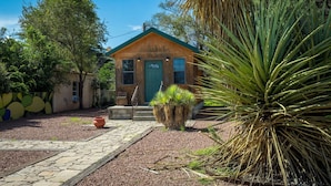 Una pequeña casa de madera con puerta verde, rodeada de un jardín con diversas plantas y un sendero de piedra que conduce a la entrada.