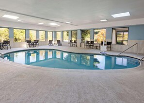 Une piscine intérieure à l’eau d’un bleu cristallin, entourée de chaises longues et de tables, et dotée de grandes fenêtres offrant une vue sur les arbres.