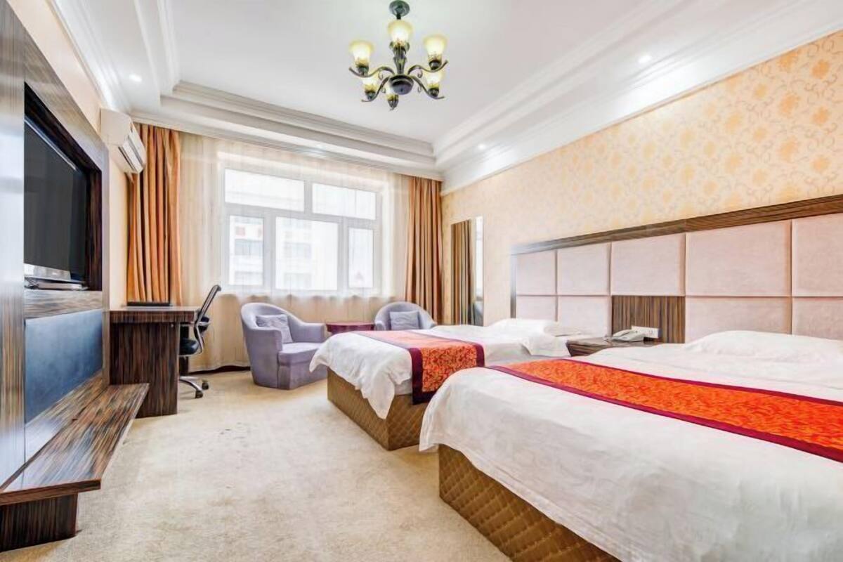 Quarto de hotel com duas camas, uma escrivaninha, uma cadeira, televisão e uma janela grande.
