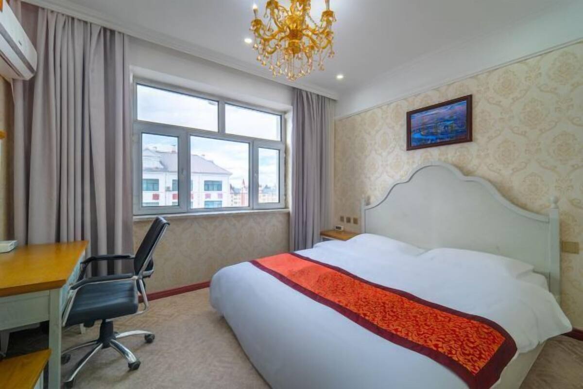 Quarto de hotel com uma cama grande, uma mesa de trabalho e uma cadeira. Há uma janela com cortinas, um candelabro e um quadro na parede.