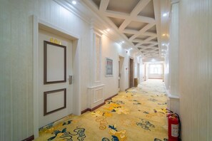 Un couloir d’hôtel avec un tapis à motifs, des murs blancs et des portes portant des étiquettes.