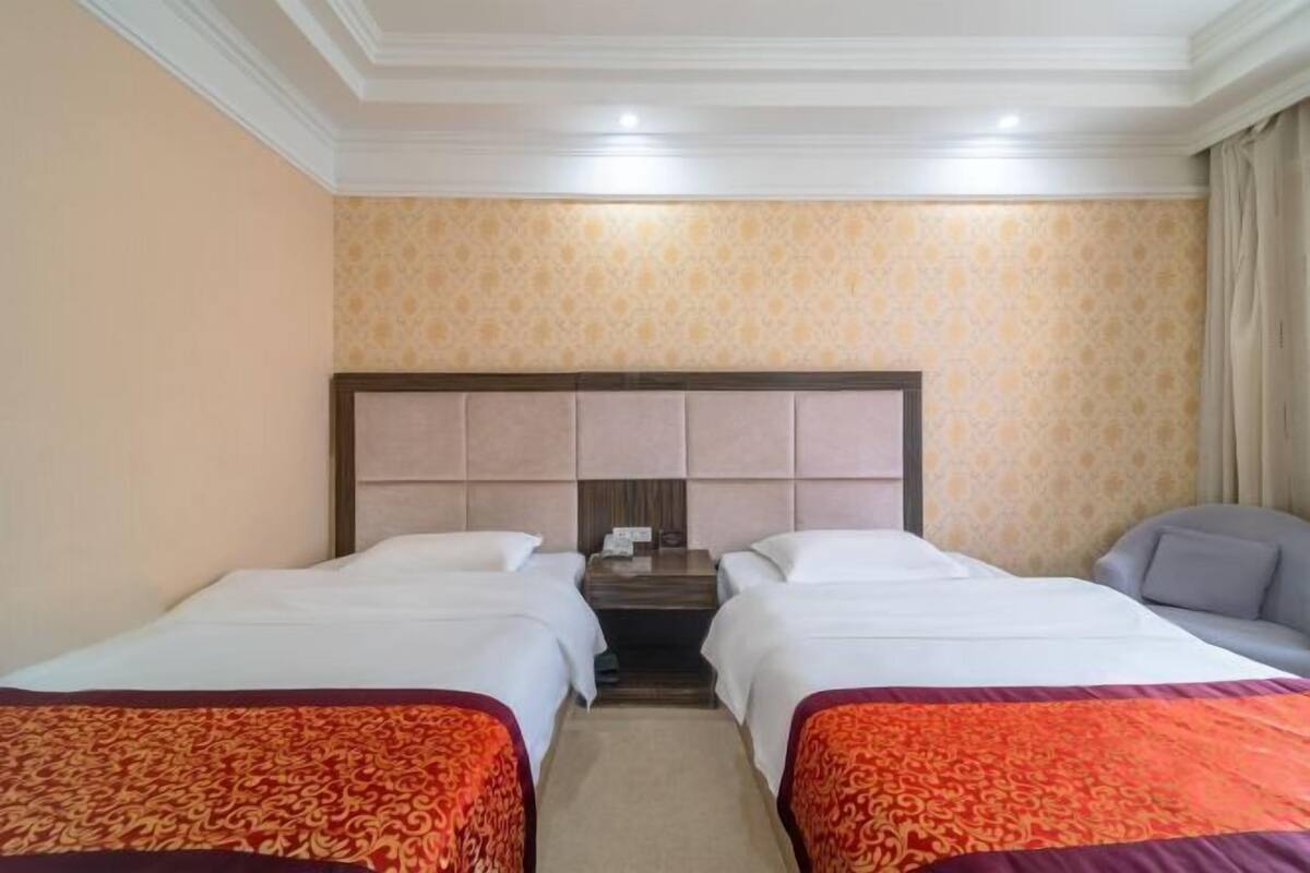 Quarto de hotel com duas camas de solteiro, uma cabeceira de madeira, uma mesa pequena e um sofá cinzento.
