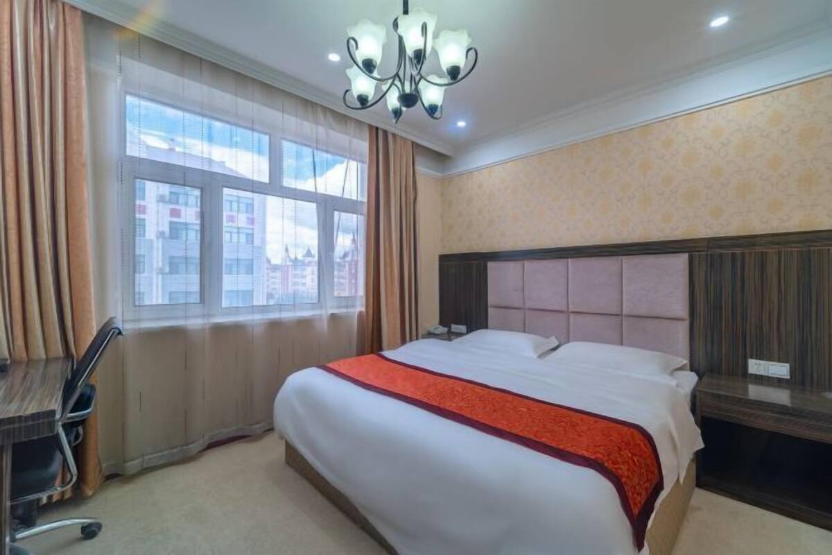Quarto de hotel com uma cama grande, uma escrivaninha e uma cadeira. Há uma janela com cortinas e vista para edifícios.