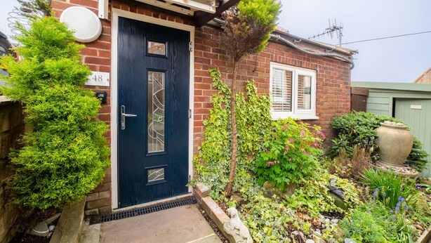 A brick house with a dark blue door, a small garden with various plants, and a number '48' sign on the wall. - Charming guesthouse for two with private parking and en-suite (North Yorkshire)